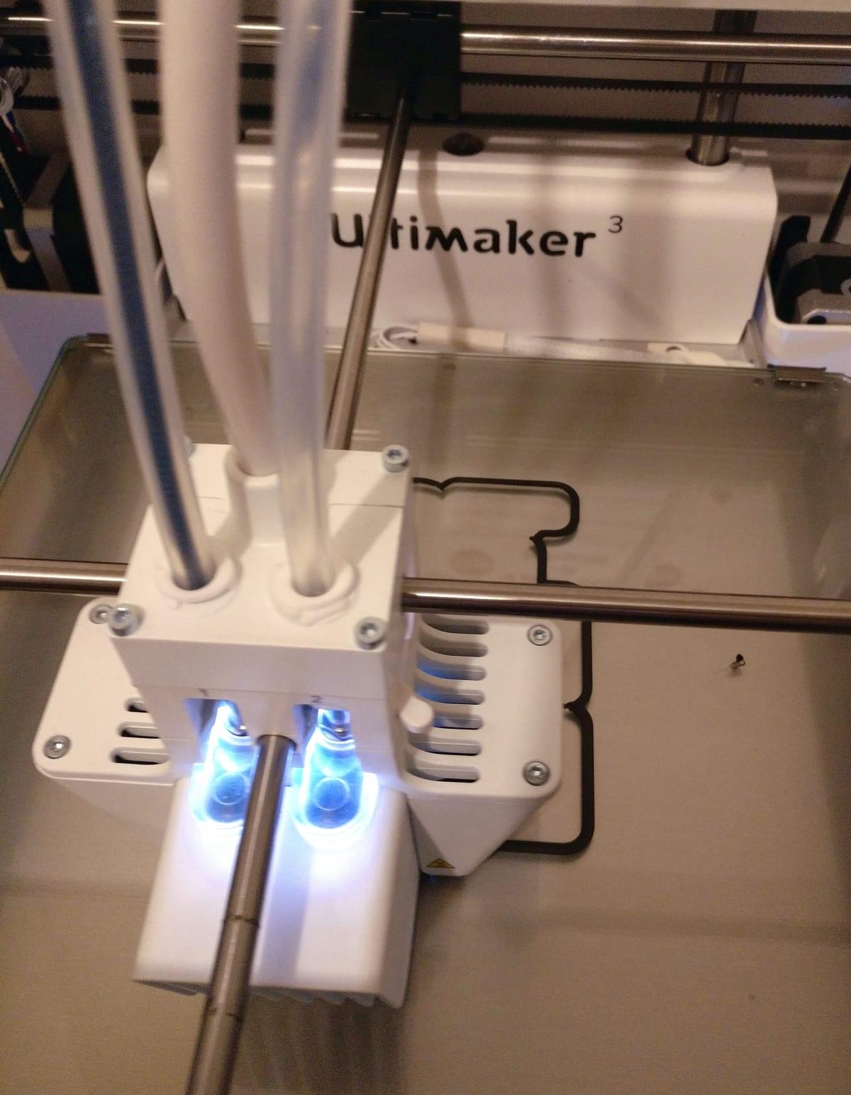 Close-up of the dual-nozzle toolhead of an Ultimaker 3 FDM printer sitting over the glass build plate at the start of a print