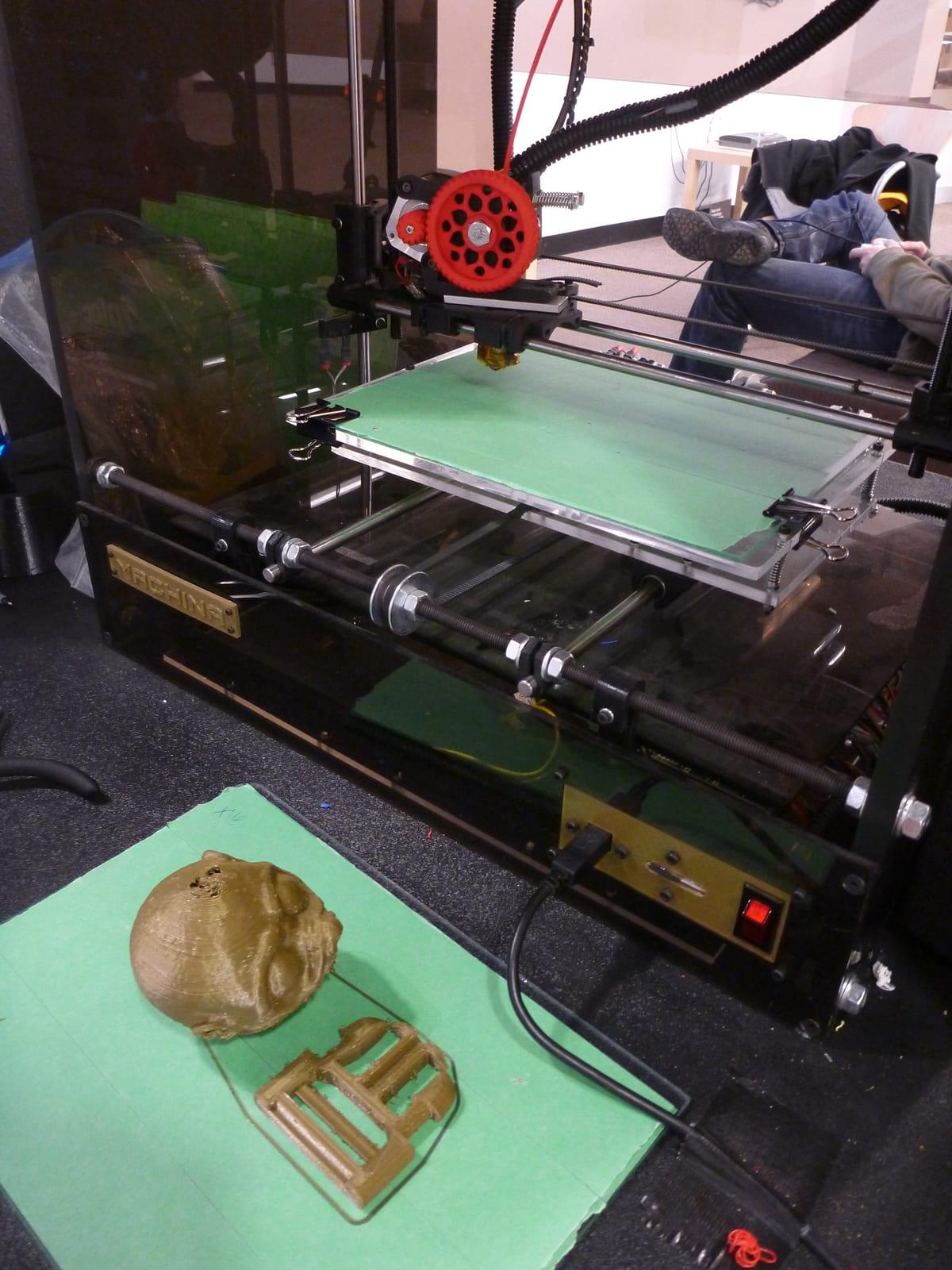 An open-frame 3D printer on a workshop bench with two freshly printed gold-coloured parts laid on a green cutting mat in front of it, a skull and a rectangular frame, both showing visible layer lines