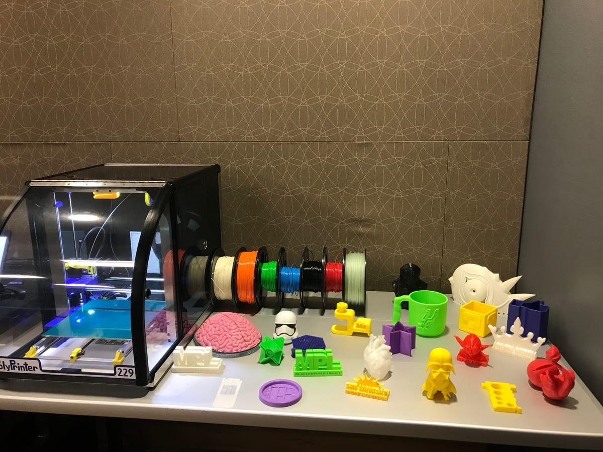 An enclosed desktop 3D printer beside filament spools and a table of small printed samples in a public library