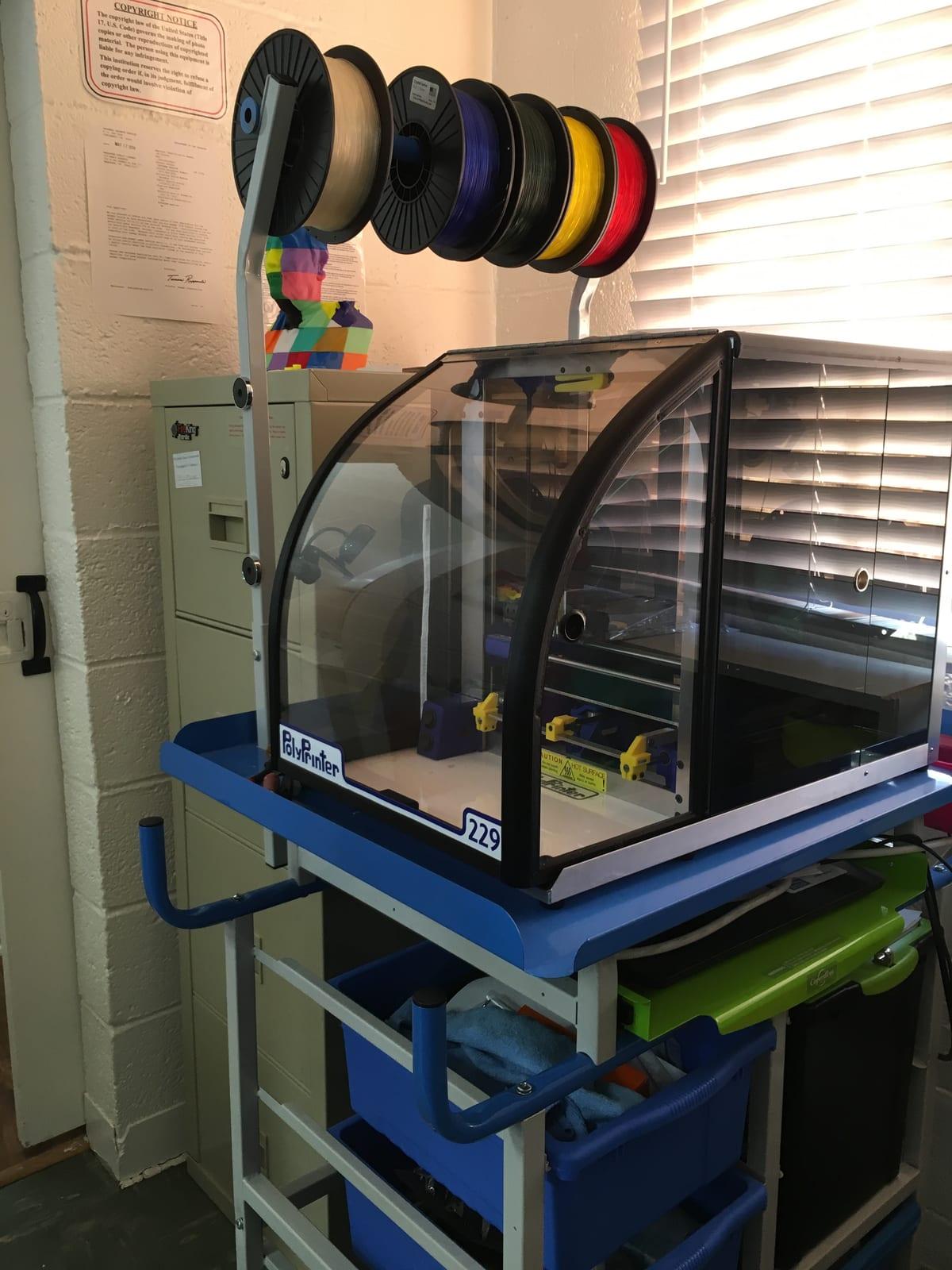 Enclosed desktop 3D printer on a rolling cart in a public library, with six spools of filament on a rod above it and a copyright notice posted on the wall