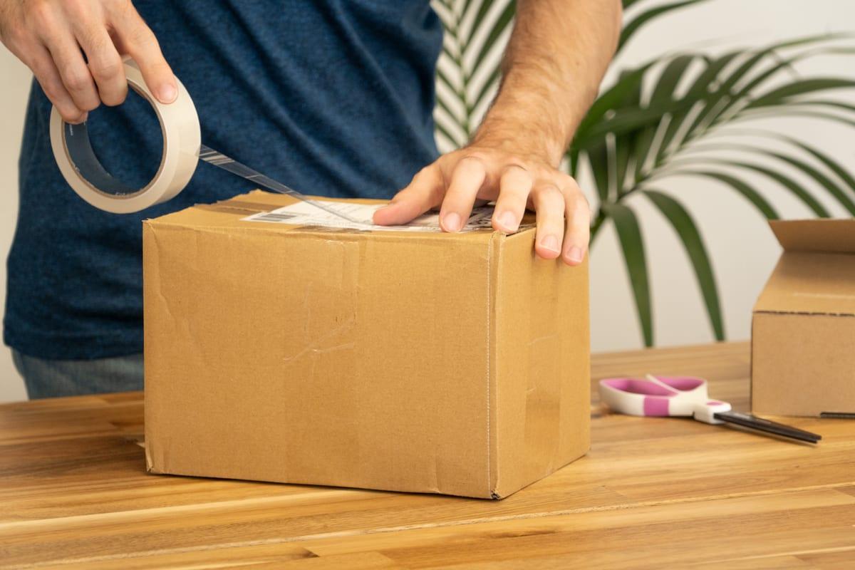 Person taping a shipping label onto a small cardboard parcel on a wooden table