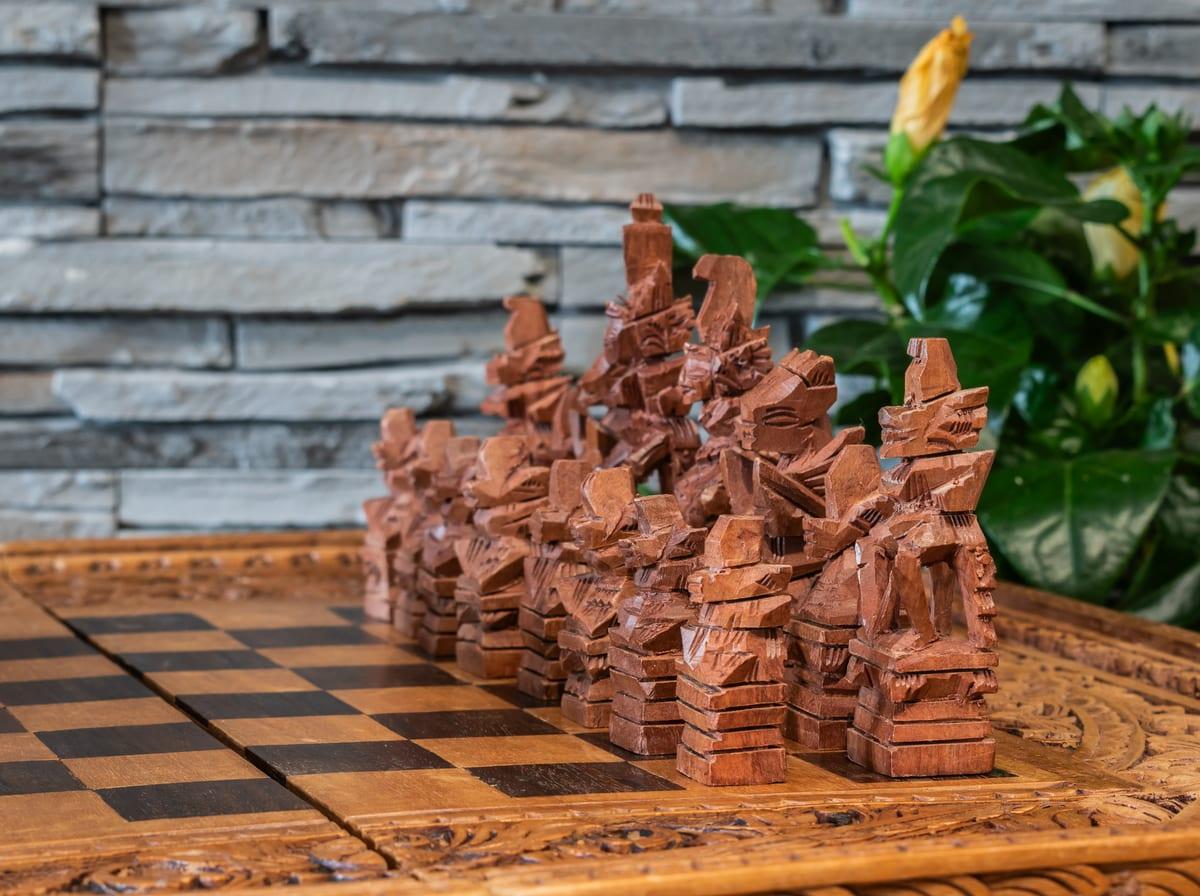 A row of hand-carved wooden chess pieces in an unconventional, non-Staunton design, standing on a carved wooden board