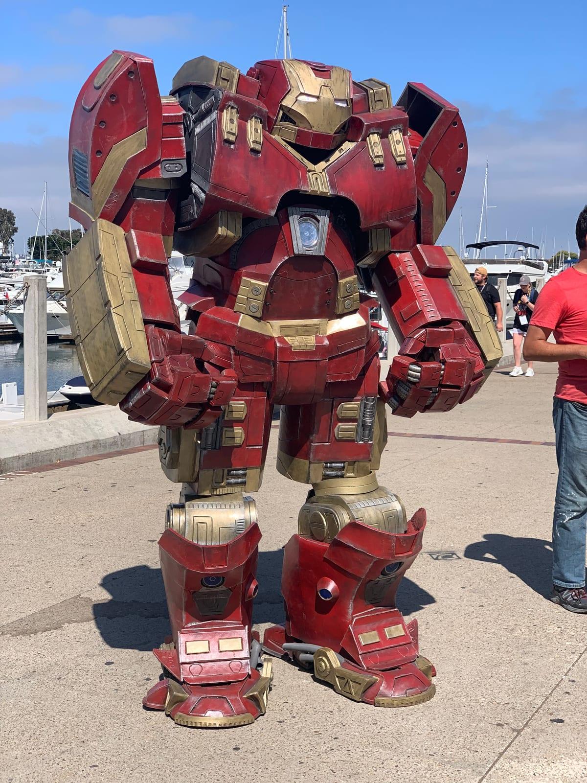A very large red and gold Hulkbuster armour cosplay suit with oversized shoulder, arm and chest plates, photographed outdoors on a dockside