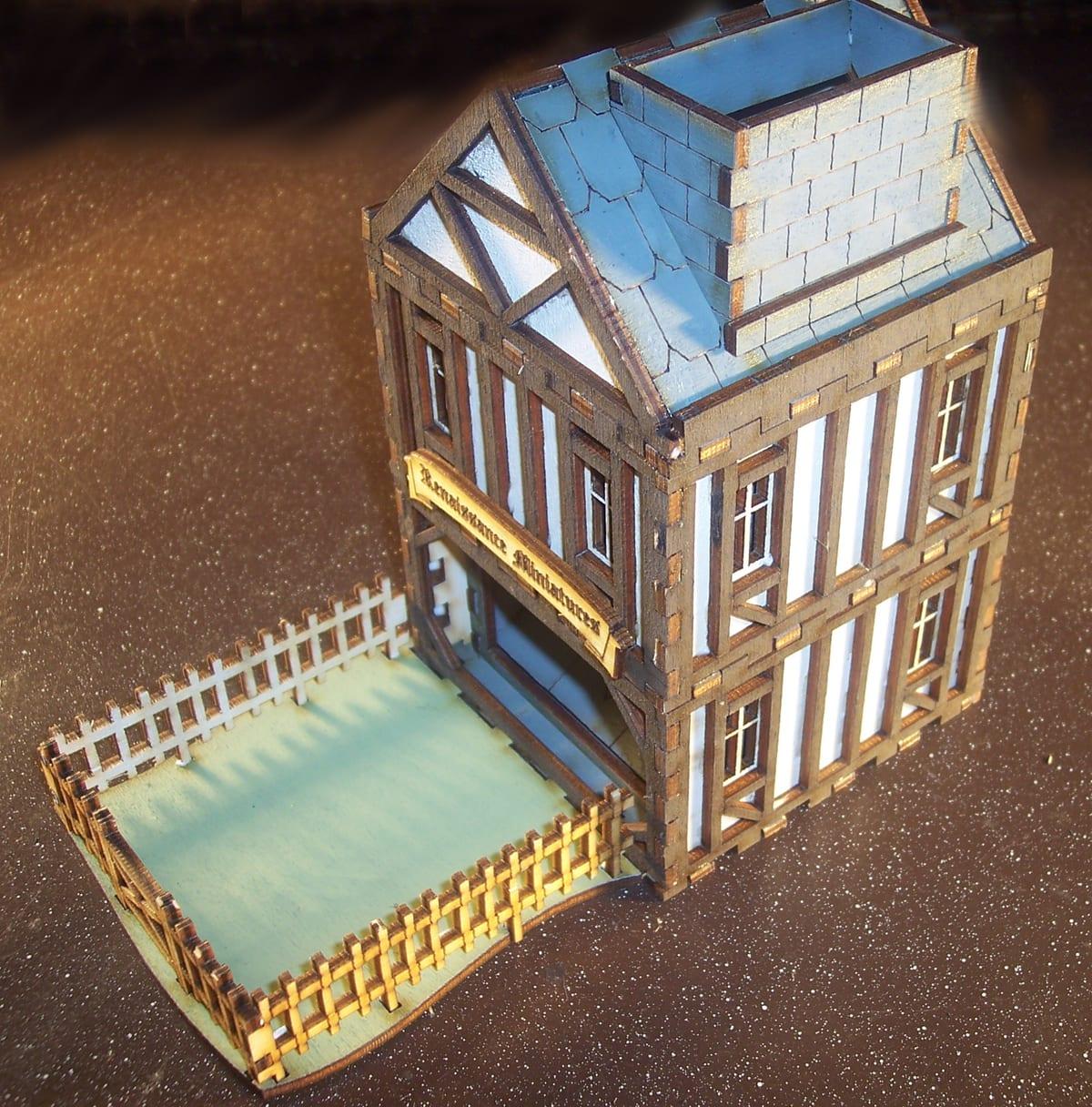 A dice tower built to look like a medieval half-timbered shop, assembled from laser-cut wooden parts, with a fenced catch tray in front