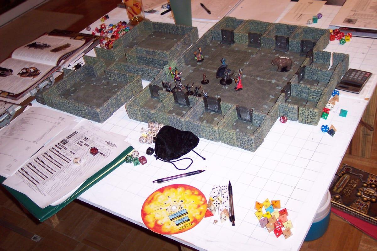 Miniatures inside a modular dungeon built from stone-effect wall and floor tiles, set on a gridded tablecloth surrounded by dice, rulebooks and character sheets