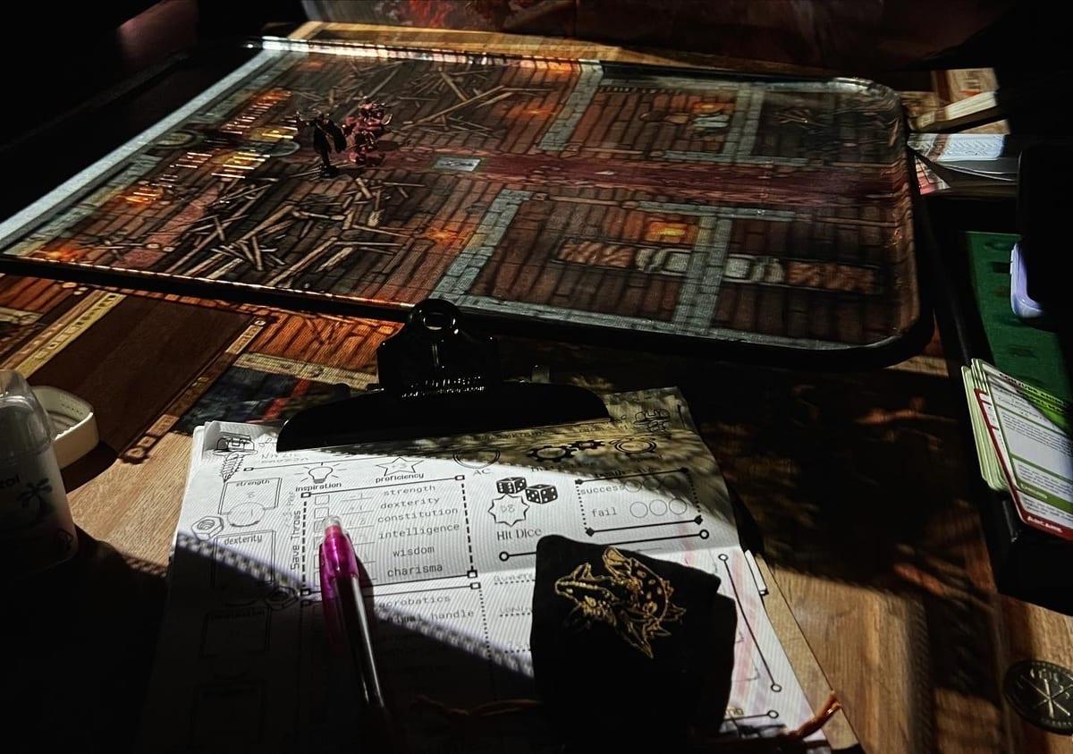 A dimly lit Dungeons and Dragons table with a laminated dungeon battle map, a single miniature on it, a character sheet under a clipboard, a pen and a dice bag