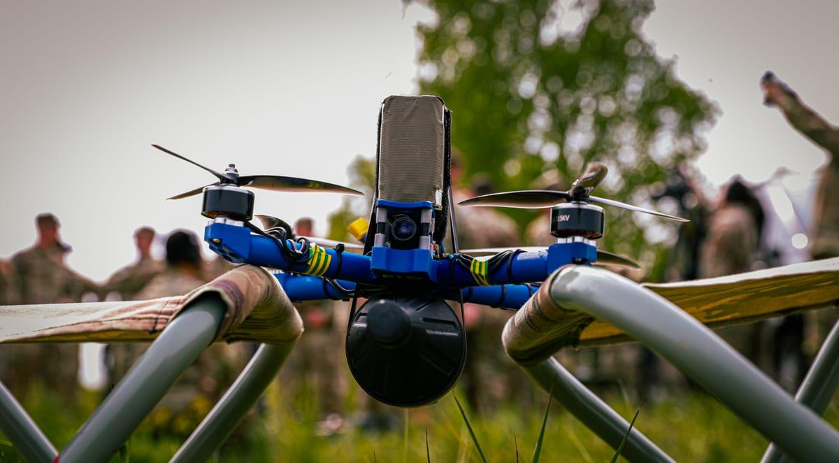 An experimental quadcopter with a blue printed airframe and a camera at the nose, resting on a folding cot frame in a field with soldiers blurred in the background