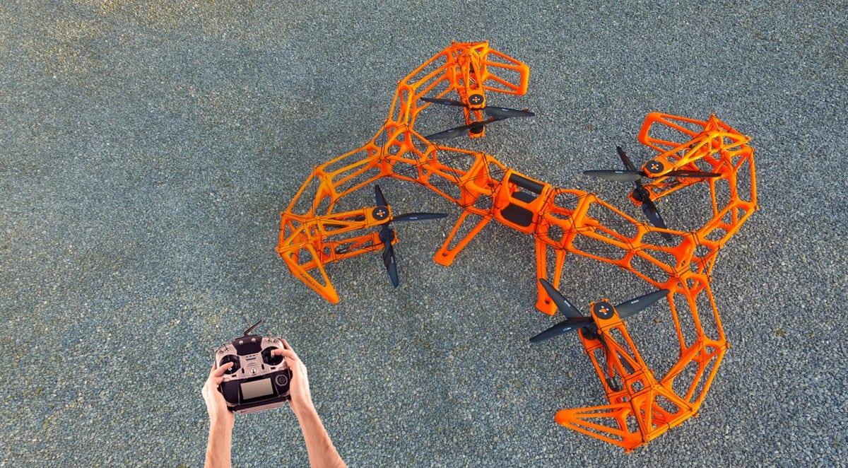 A large orange 3D printed lattice-frame multirotor drone standing on tarmac, with a pilot holding a transmitter in the foreground for scale