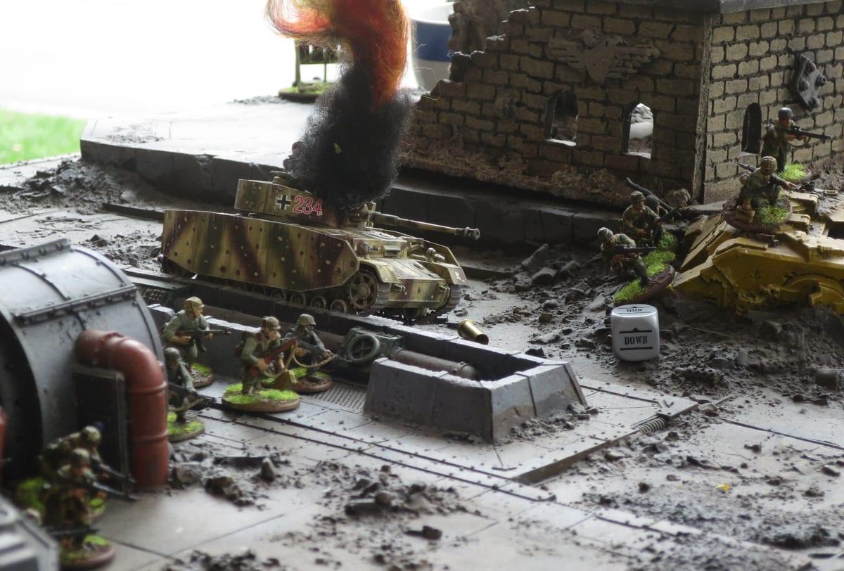 Close-up of a WWII wargaming table: a camouflaged tank on a paved street beside a shell-damaged brick building, with infantry miniatures, rubble and a knocked-out vehicle