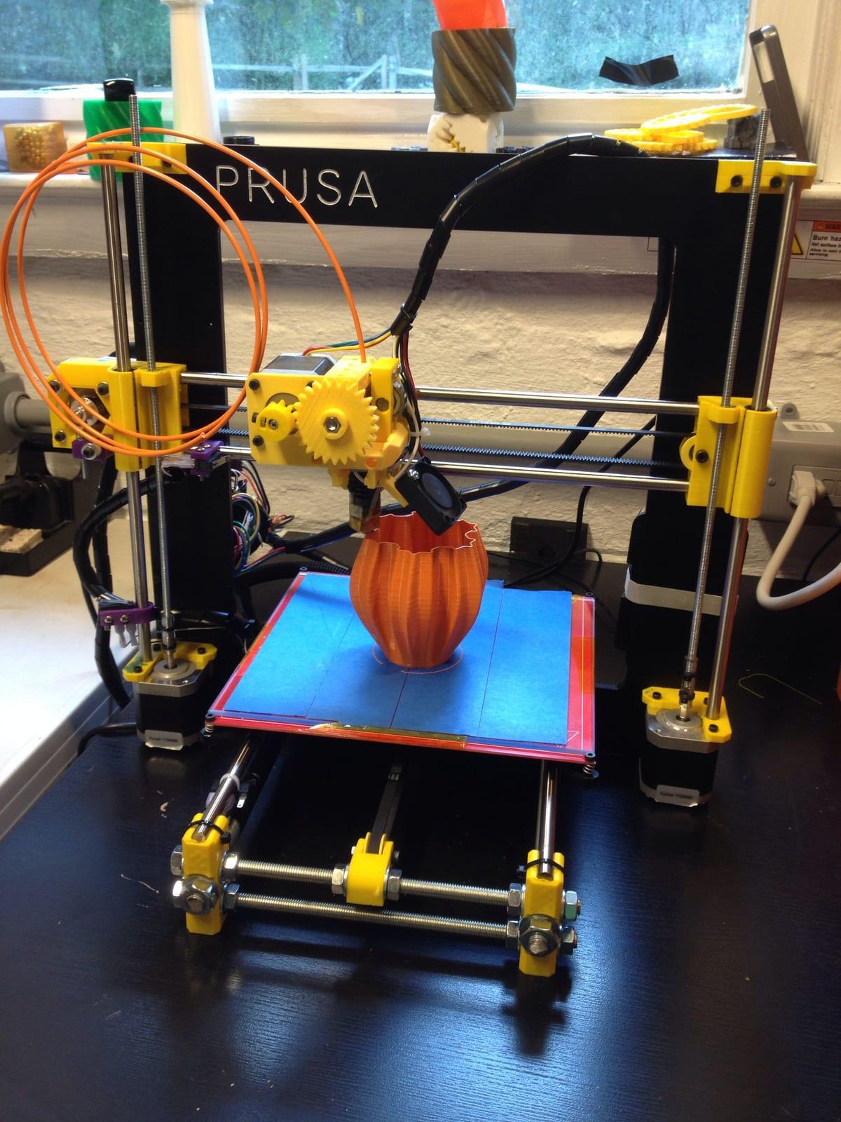 A RepRap-style Prusa i3 3D printer mid-print, extruding a tall thin-walled vase on the heated bed