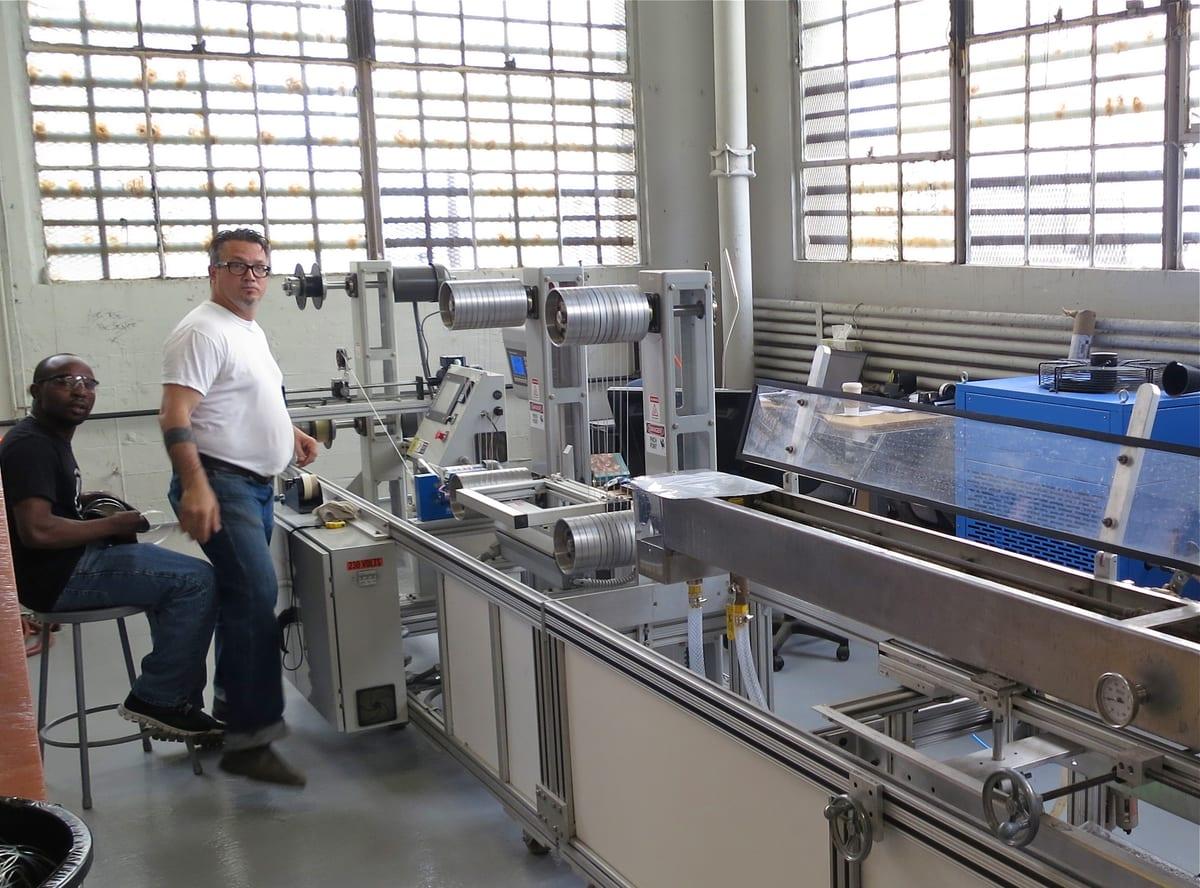 Two workers standing beside an industrial filament extrusion line in a factory, with a long cooling trough running down the right of the frame and puller rollers and a winder at the far end