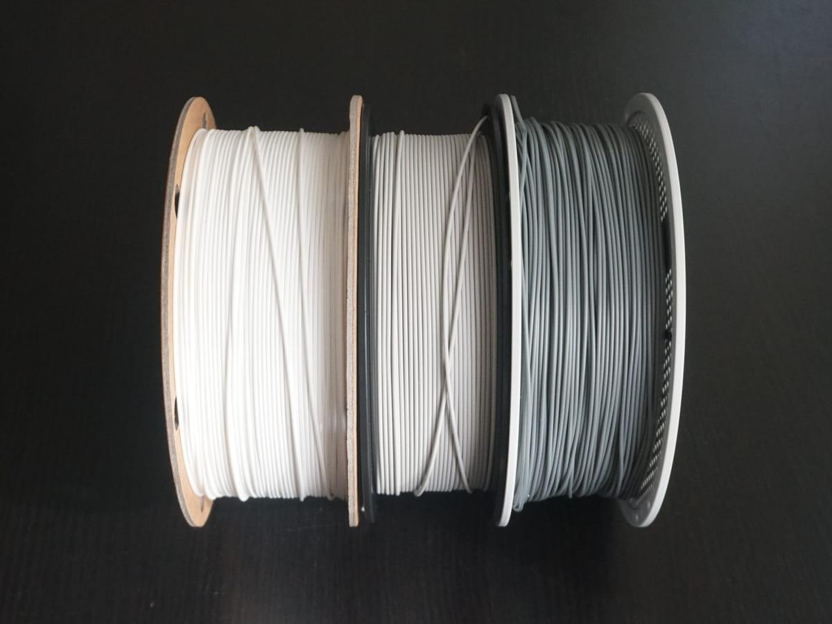 Three one-kilogram filament coils side by side: one on a cardboard reel, one on a black plastic reel, and one refill coil on a reusable white reel