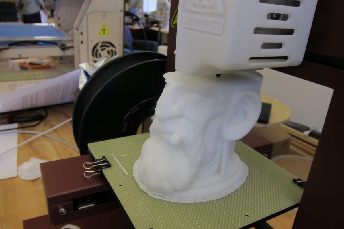 A white printed bearded head part-finished on a green build plate, the print head sitting directly above it and a spool of filament feeding in from behind