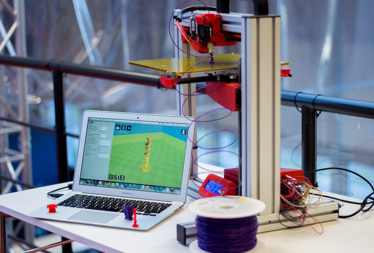 A red-and-aluminium desktop FDM 3D printer on a white bench next to an open laptop showing slicer software, a spool of purple filament and three small finished printed figures
