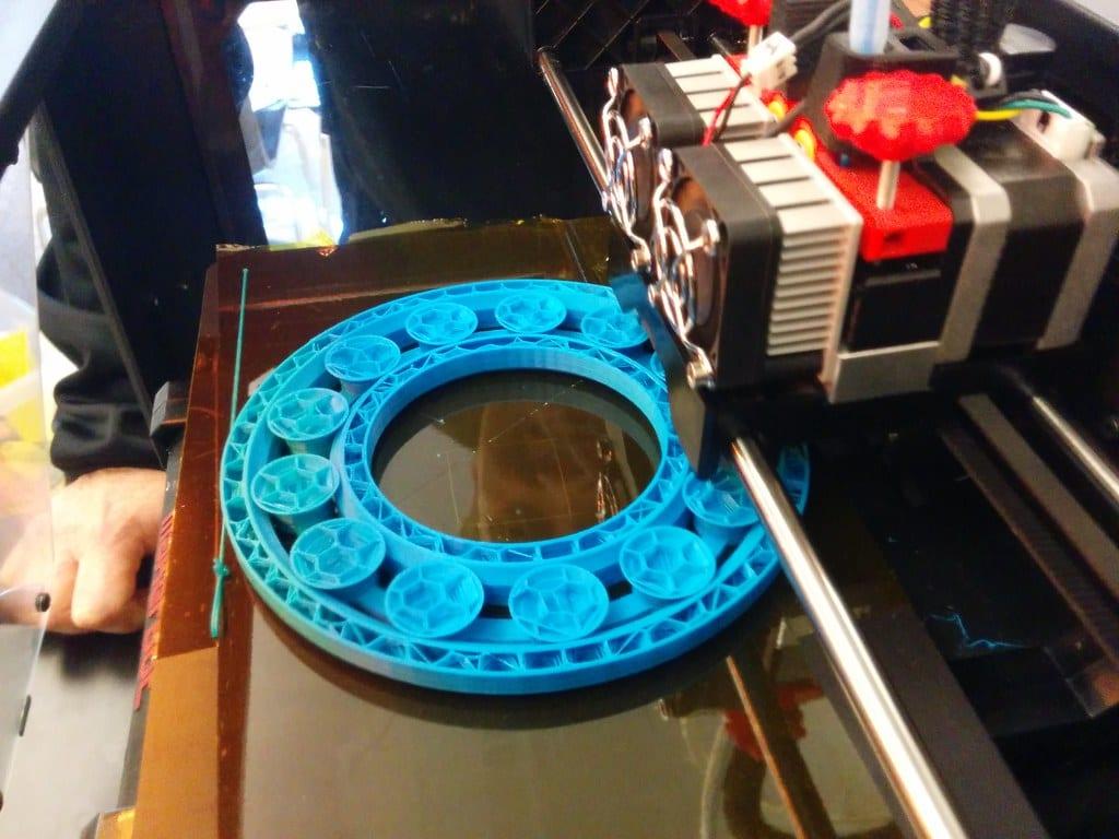 A large blue 3D printed slewing bearing sitting on a printer bed, its rollers visible in open pockets between the inner and outer races