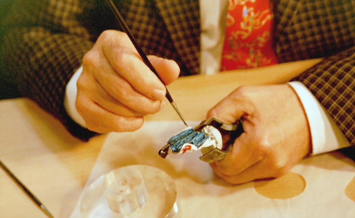 Close-up of two hands holding a small figurine and a fine brush, painting detail onto it at a table
