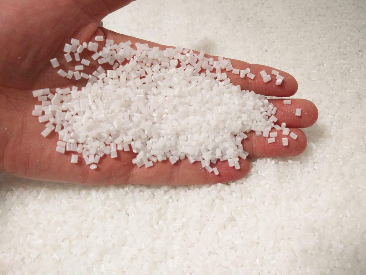 A handful of small opaque white ABS polymer granules held over a bin full of the same pellets