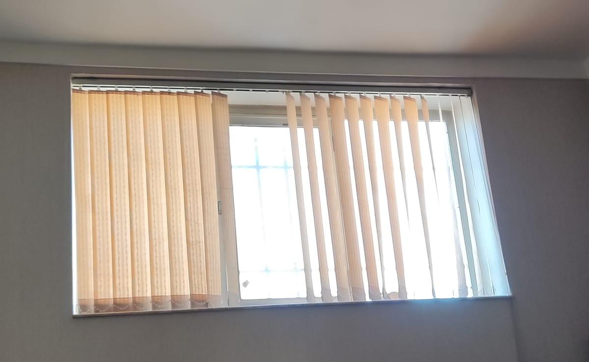 Window fitted with vertical fabric blind vanes hanging from a headrail, partly drawn to one side