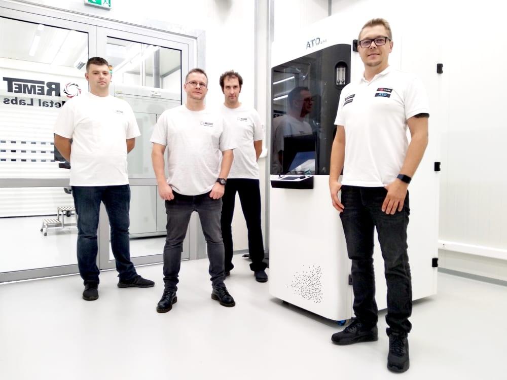 Four engineers in white shirts standing beside a tall white metal powder atomising unit inside a clean industrial metal 3D printing laboratory