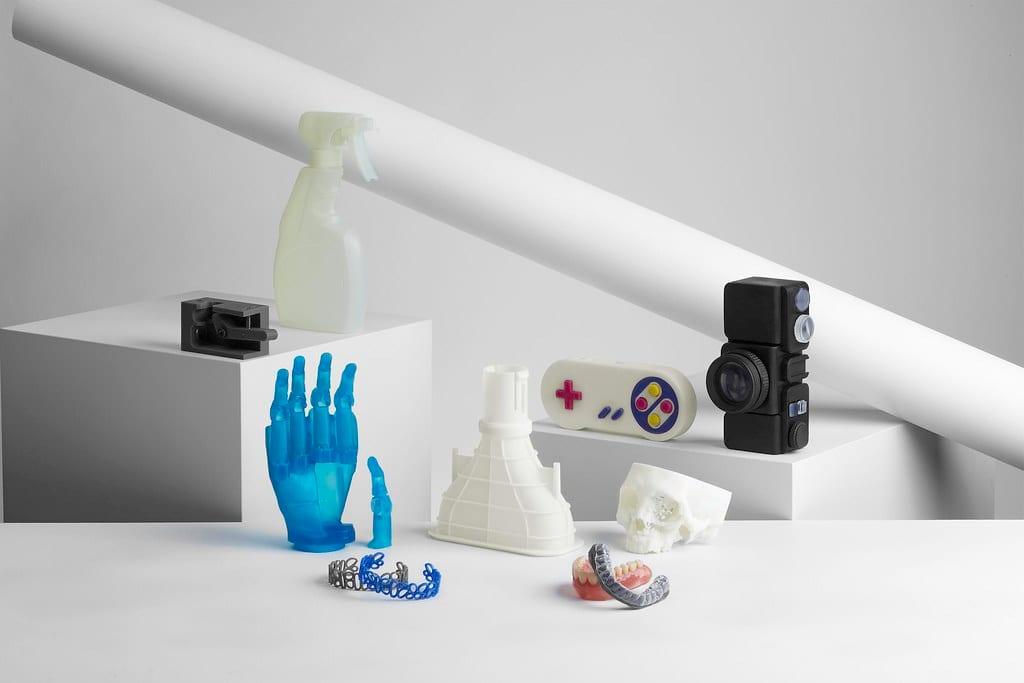 An assortment of resin 3D printed parts arranged on white plinths: a blue prosthetic hand, a translucent spray bottle, a games controller, a camera body, a skull section, dental models and a chain bracelet