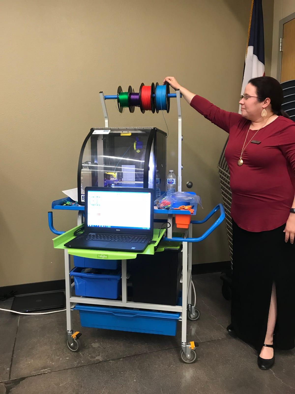 A library staff member standing beside a wheeled cart carrying one enclosed 3D printer, a laptop and four spools of coloured filament on a rail above it
