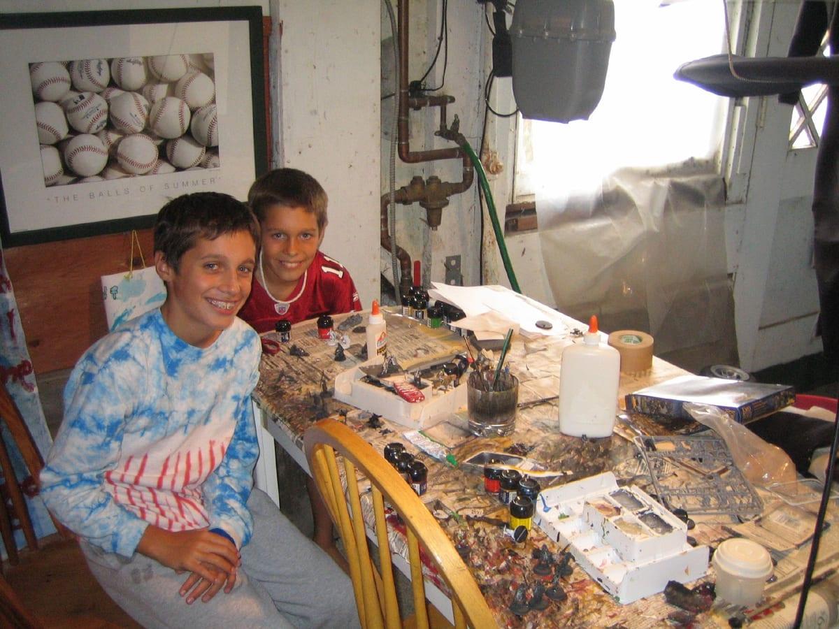 Two young hobbyists at a cluttered painting bench covered in newspaper, pots of acrylic paint, brushes, glue and part-painted tabletop miniatures on trays