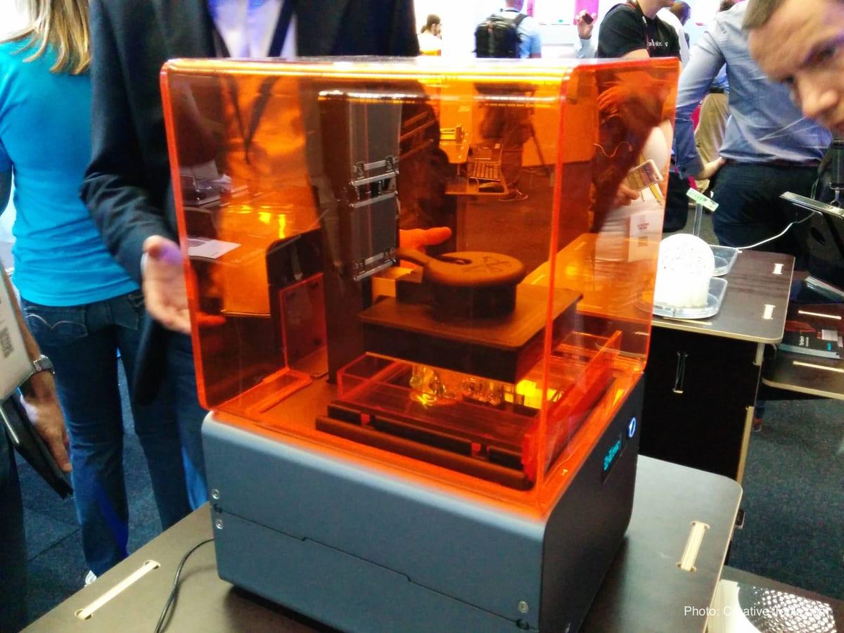A desktop SLA resin 3D printer with its orange cover, shown on an exhibition stand