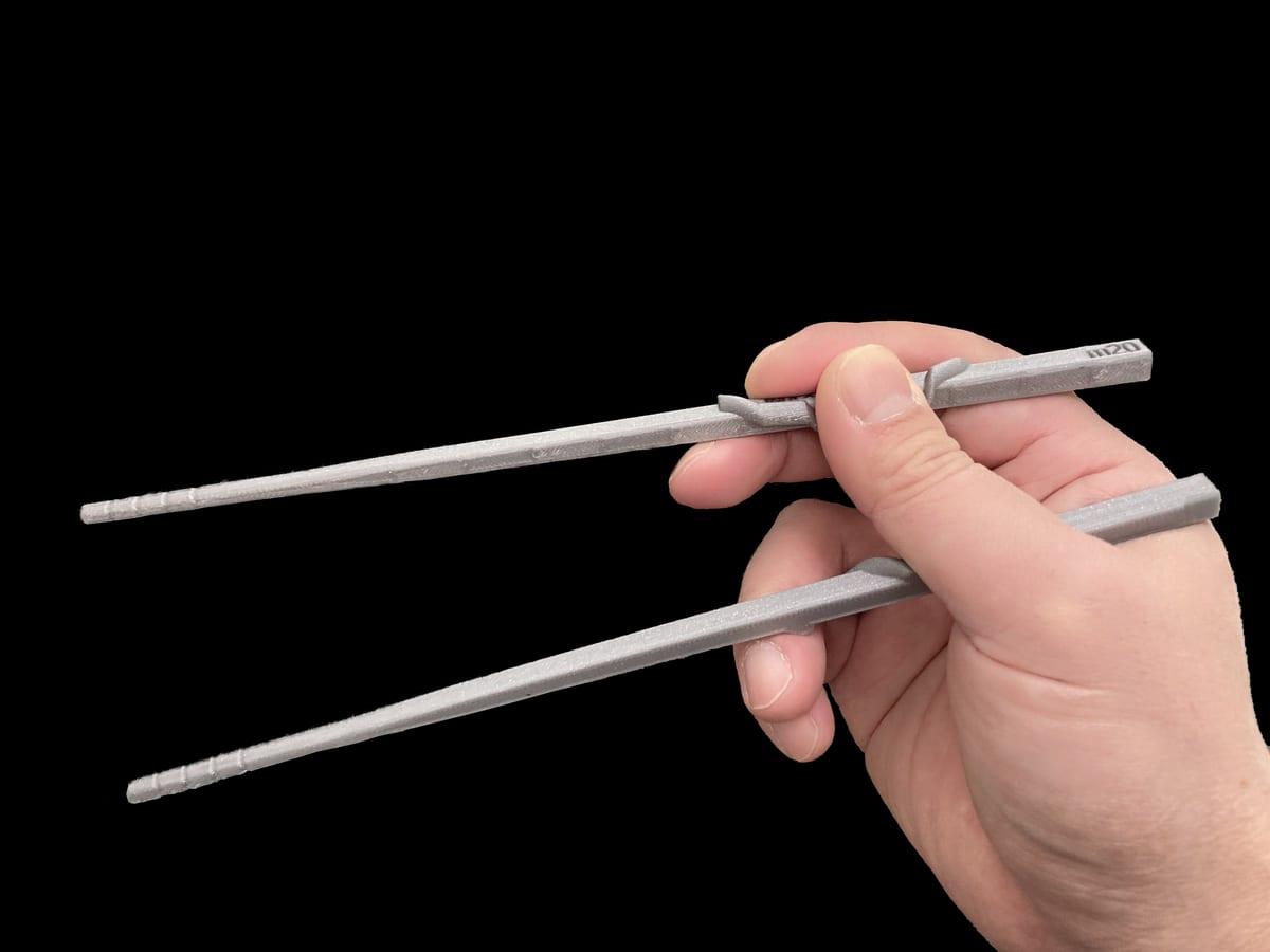 A hand holding a pair of grey 3D printed ergonomic chopsticks with a moulded finger notch, against a black background