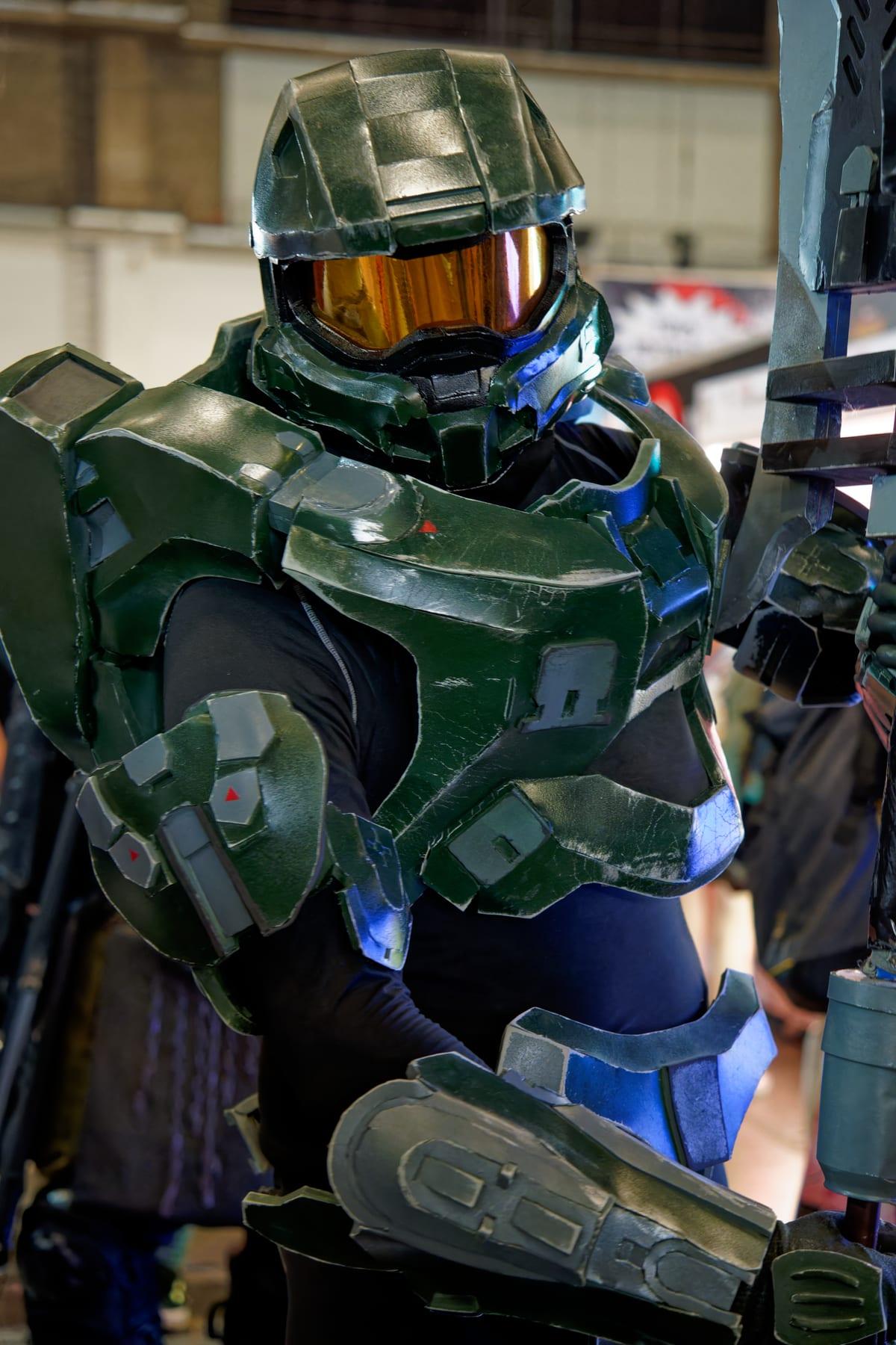 A cosplayer in a green sci-fi armoured suit with a full helmet and gold reflective visor, wearing layered shoulder and chest plates