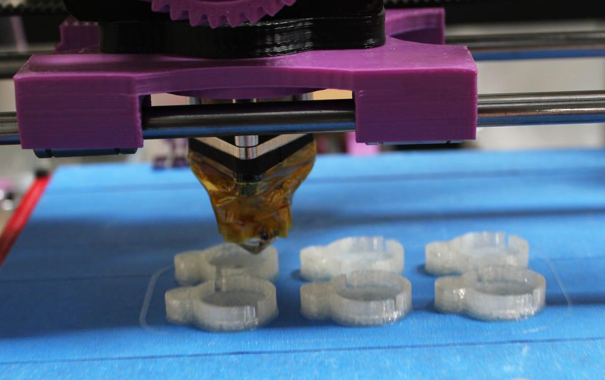 Close-up of a 3D printer print head mid-print, showing the nozzle and a heater block crusted with oozed filament, laying down small translucent rings on a blue-taped bed
