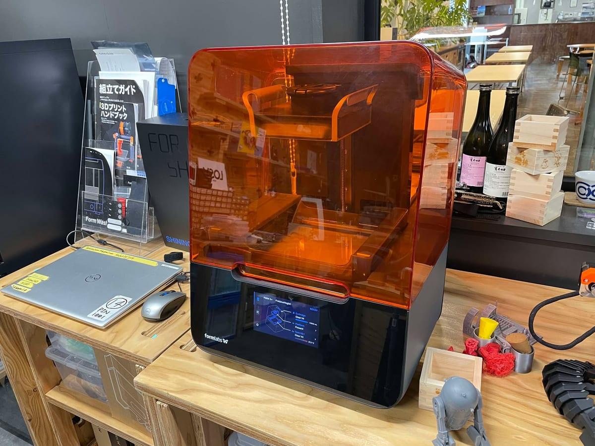 A desktop resin 3D printer with an orange hood standing on a workbench in a fab lab, with small printed parts and a grey printed figure on the bench beside it