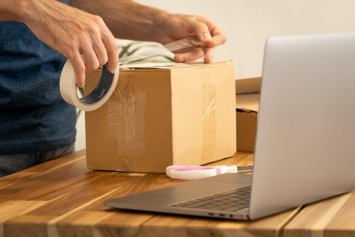 Person sealing a cardboard parcel with tape beside an open laptop used to book the shipping
