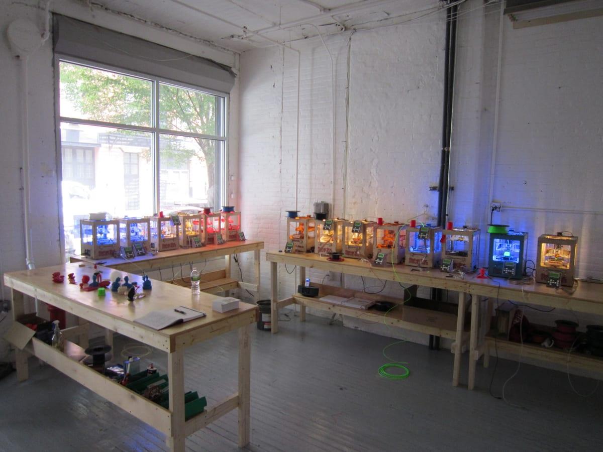 Two workbenches of wooden-framed desktop 3D printers running in a whitewashed brick workshop