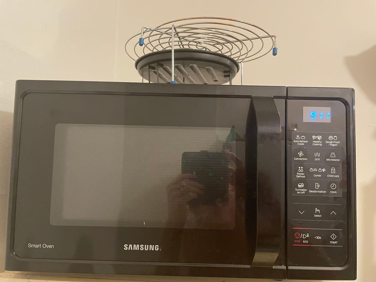 Black Samsung convection microwave oven with a moulded door pull, perforated door screen and a metal grill rack sitting on top