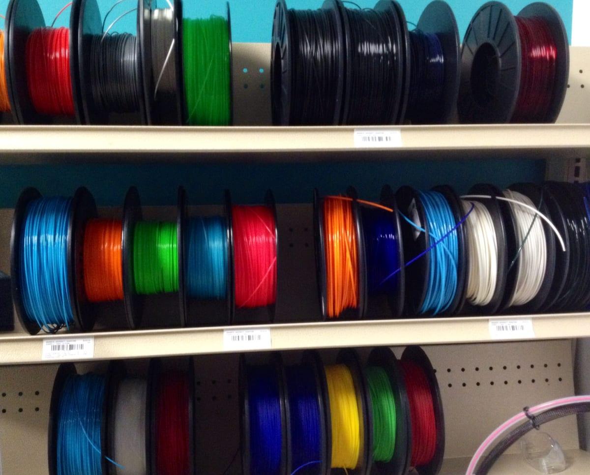 Shelves in a maker space stacked with dozens of filament spools in many different colours and materials