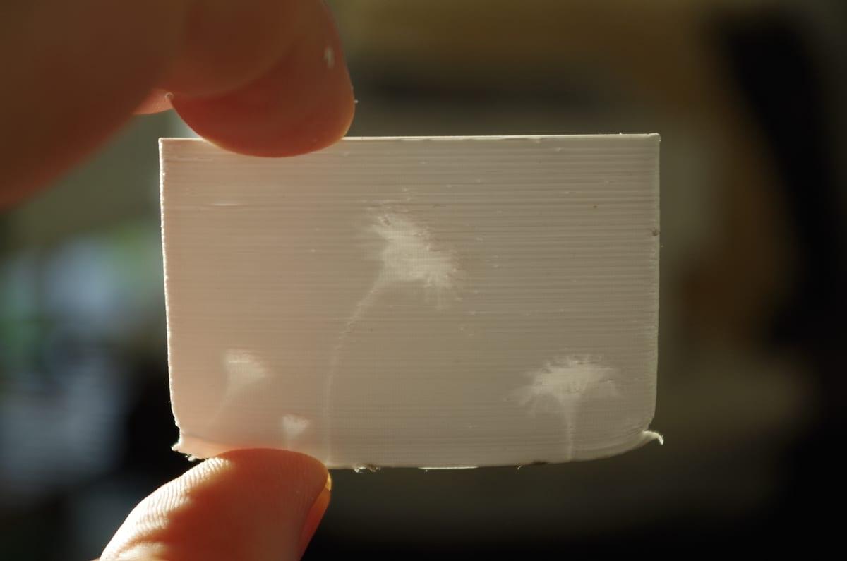 A small white FDM printed lithophane held between finger and thumb against a window, faintly showing a flower and stem through the plastic, with horizontal layer lines visible