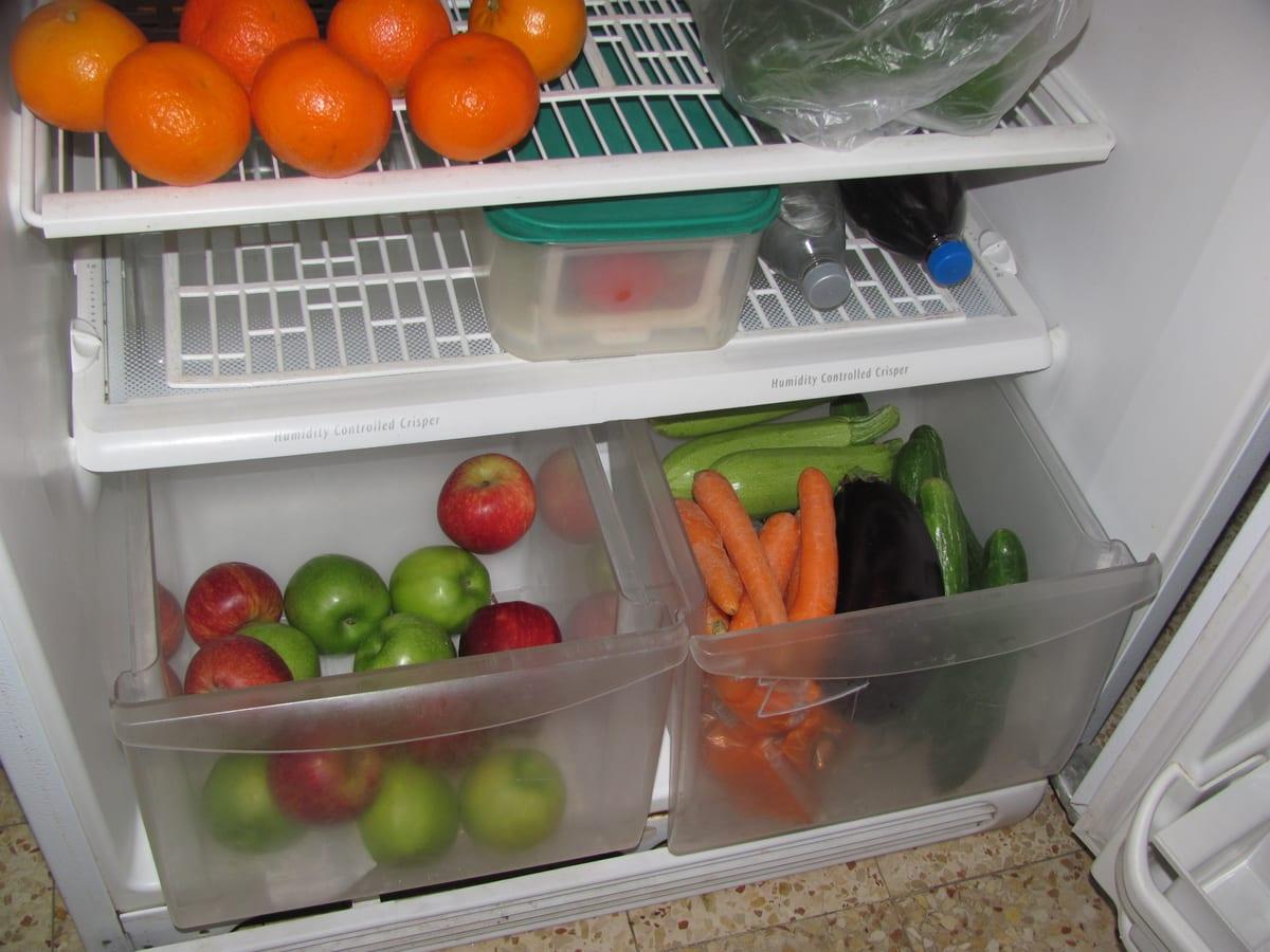 Refrigerator interior seen from above, with two translucent crisper drawers of apples and vegetables beneath a humidity-controlled crisper cover and wire shelves