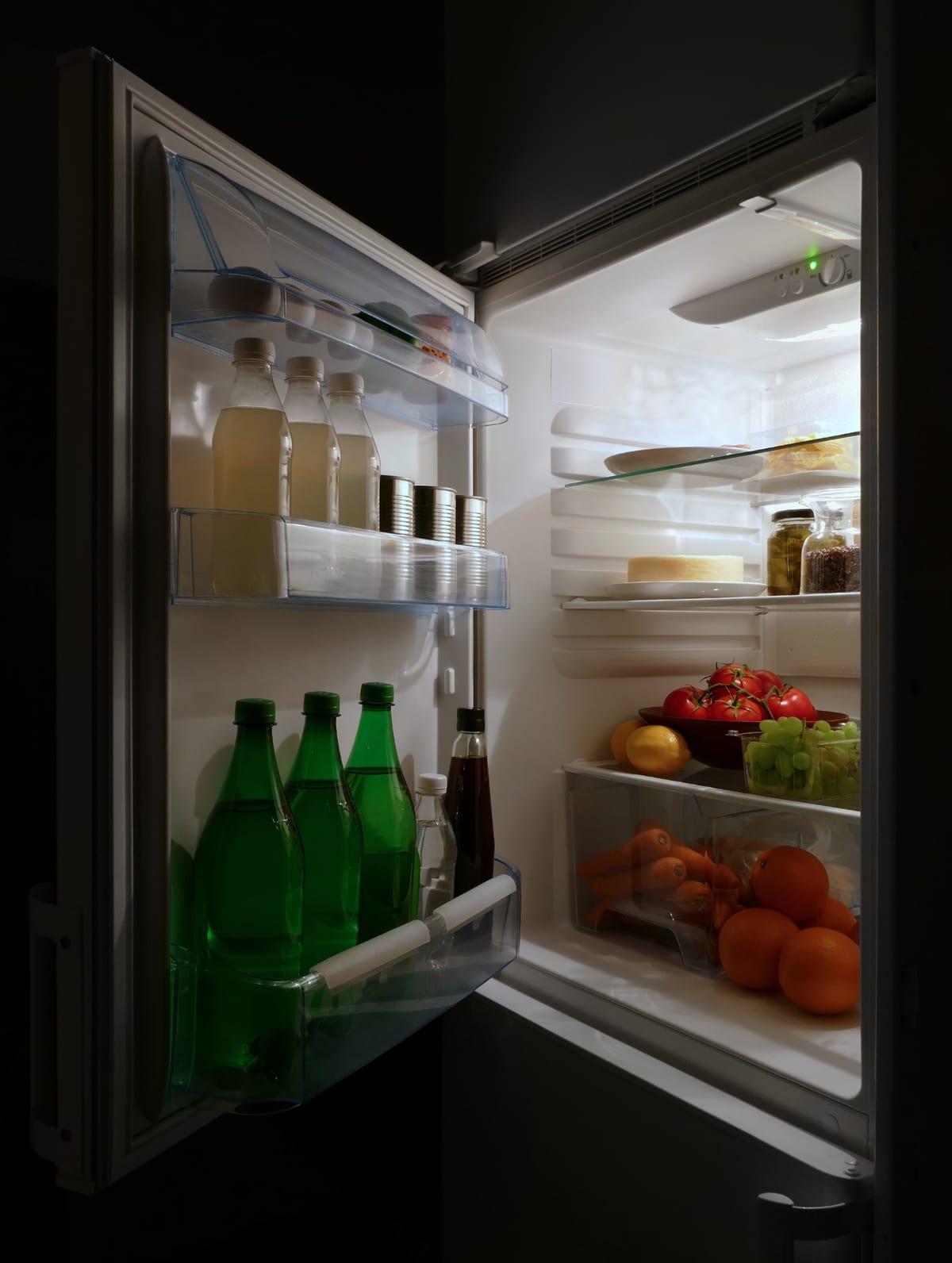 Refrigerator with the door open at night, interior shelves holding plates, a cheese and jars, a crisper drawer of carrots and oranges below, and bottles and cans in the door bins