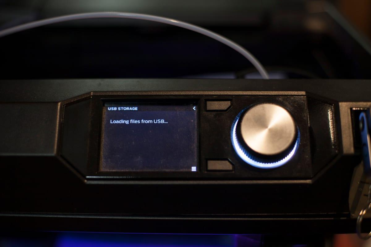 Close-up of a 3D printer's control panel and jog wheel, the small screen reading "USB STORAGE — Loading files from USB" with a memory stick plugged in beside it