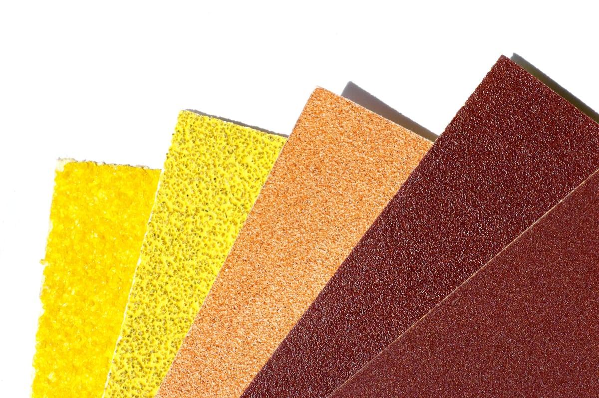 Five sheets of abrasive paper fanned out on a white background, running from a coarse open yellow grit on the left to a fine dark red grit on the right