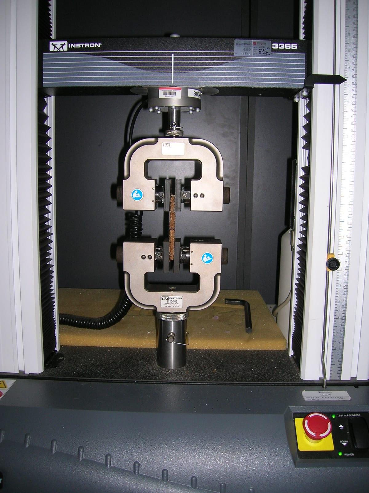 A narrow fibre-composite test specimen clamped between the upper and lower grips of an Instron tensile testing machine