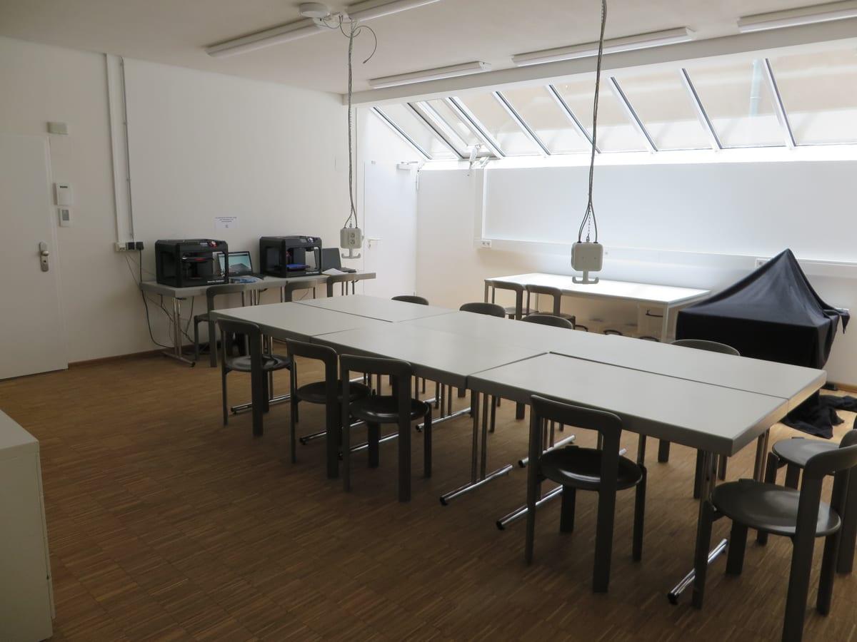 Library makerspace room with two enclosed desktop 3D printers on a side bench, facing a block of work tables and stools