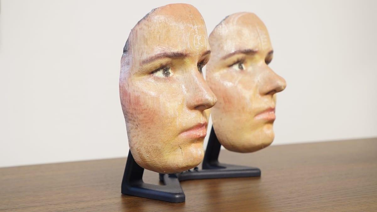 Two 3D printed human face masks on stands, hand-painted in skin tones, with faint print texture and a seam line still visible across the cheeks
