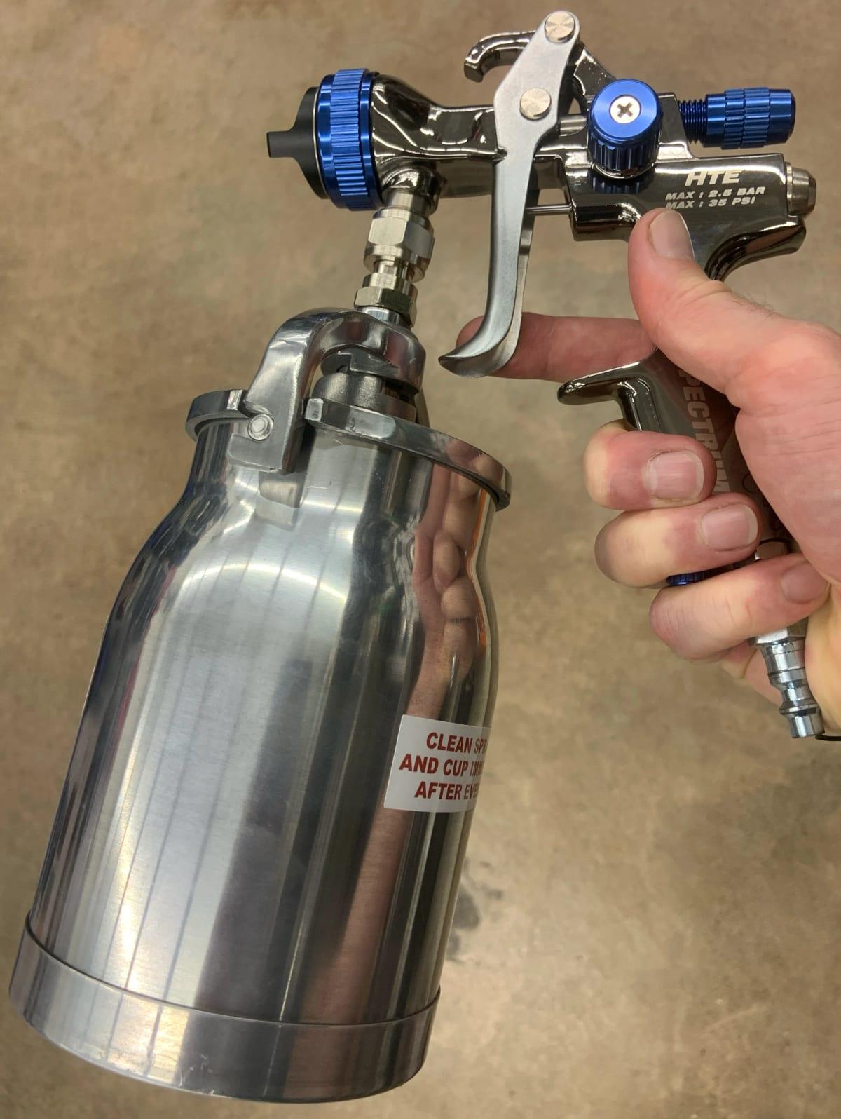 A hand holding a chrome HVLP paint spray gun with a large aluminium siphon-feed cup hanging beneath it