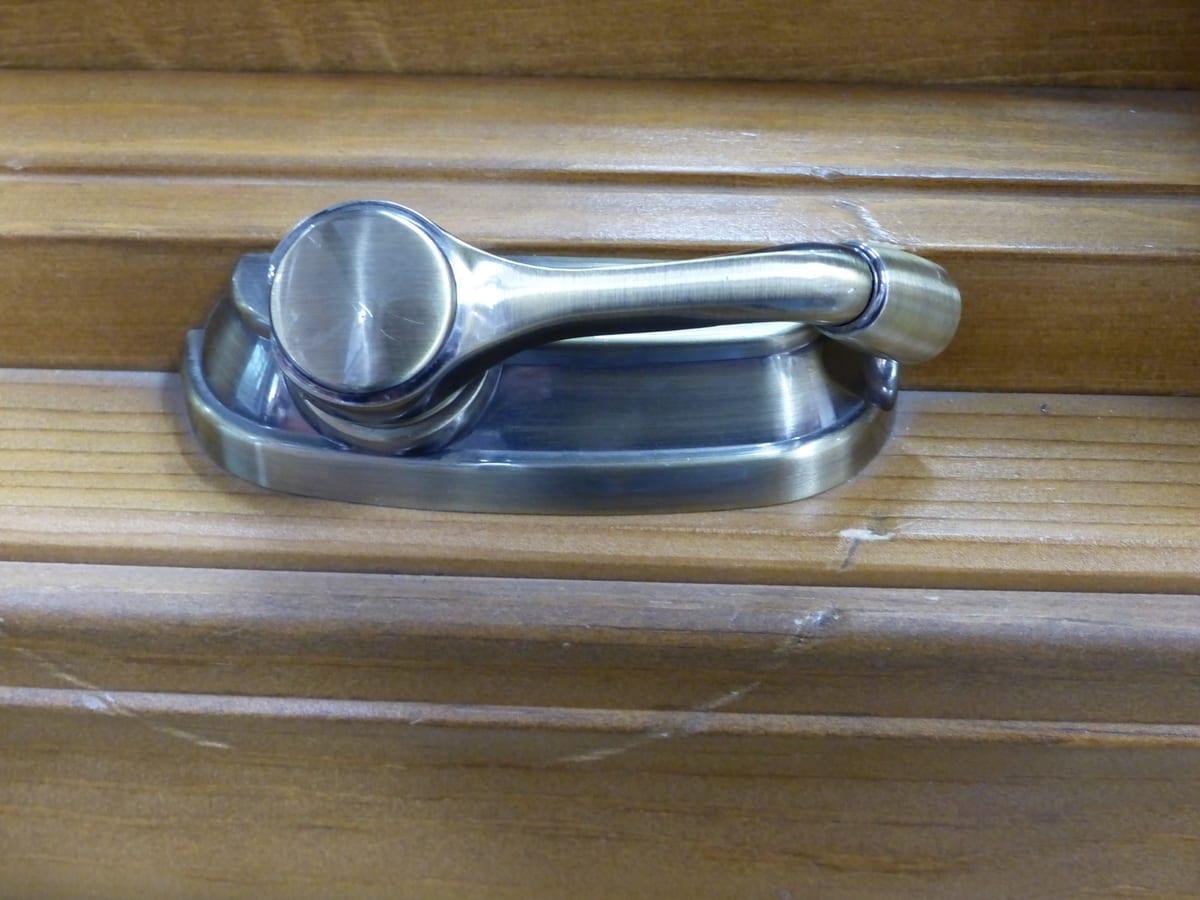 Close-up of an antique-brass folding crank handle on a wooden casement window, folded flat against its oval escutcheon plate