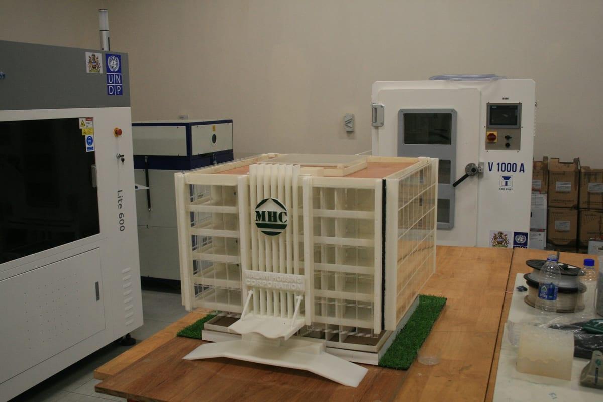 White 3D printed model of a multi-storey building beside industrial 3D printers