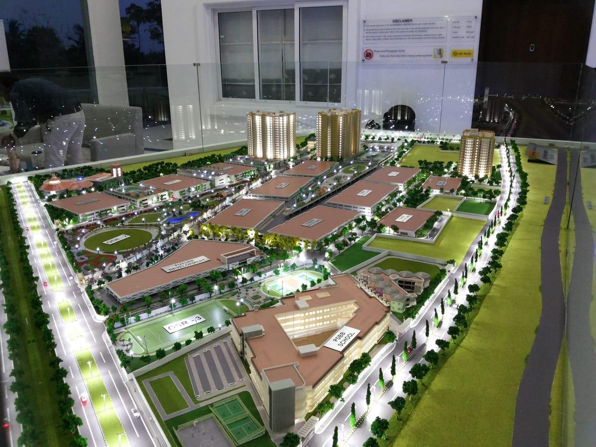 An illuminated developer masterplan model showing residential towers, low-rise blocks, roads lined with model trees, sports pitches and landscaped grounds on a large site base