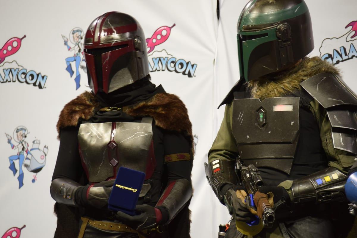 Two Mandalorian cosplayers in full helmets and metal-look armour at a convention costume contest