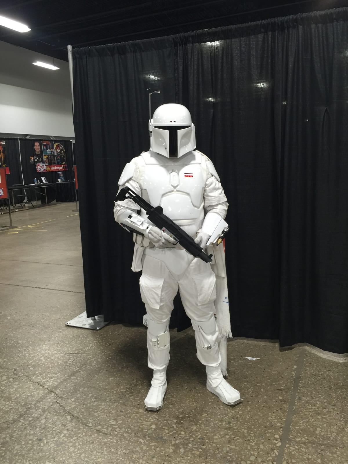 A cosplayer in full white sci-fi armor and helmet holding a prop blaster at a comic convention