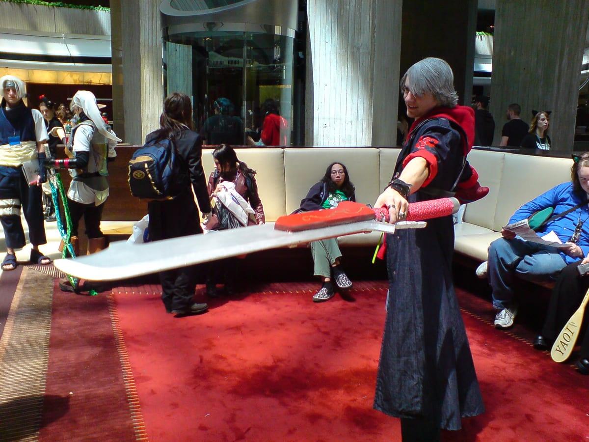 A cosplayer in a long coat holding an oversized prop sword horizontally across a convention lobby, the blade roughly as long as they are tall